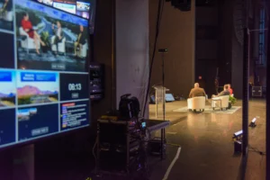 View from backstage showing two people seated on a stage in conversation, with a monitor displaying video feeds in the foreground.