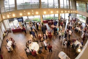 Large group of people socializing in a spacious, modern event hall with floor-to-ceiling windows, round tables, and buffet tables along the walls.