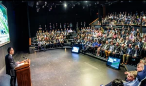 A speaker addresses a large audience seated in a tiered auditorium, with presentation screens visible on stage.