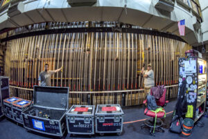 Two people stand at a theater fly system with ropes and pulleys; road cases, cables, and equipment are visible in the foreground.
