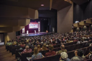 A full audience sits in a theater watching two presenters on stage with a large screen displaying images behind them.