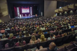 A large audience fills an auditorium, watching a presentation on stage with several speakers and a projection screen displaying information.