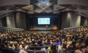 A large audience sits in an auditorium watching a speaker present slides on a stage during an event or lecture.